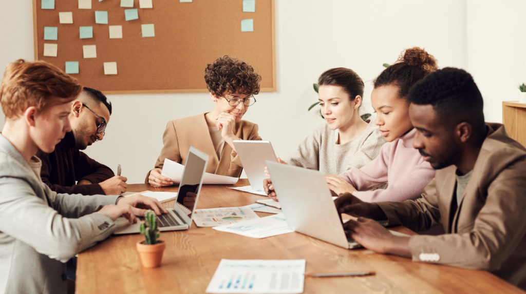 a group of diverse professionals sit at a conference table with computers and papers in discussion