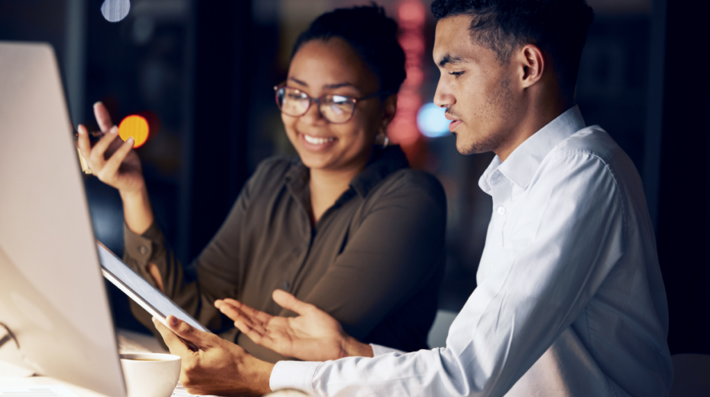 two individuals stand side by side in front of a computer, appearing to be in active conversation