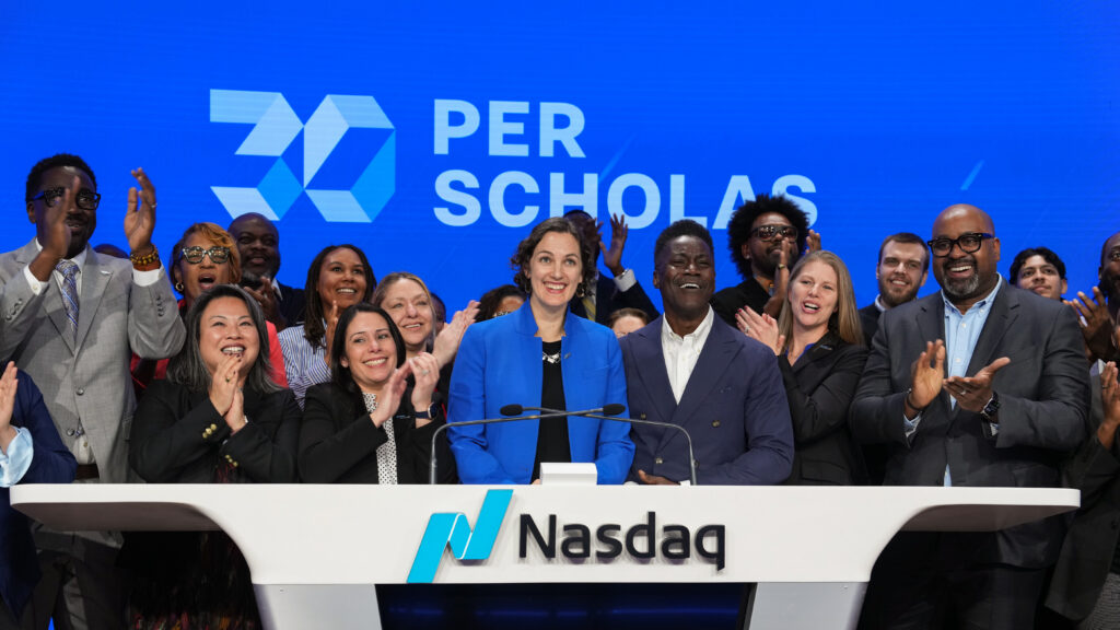 Per Scholas leadership stands in a group at the NASDAQ podium in front of a blue Per Scholas logo