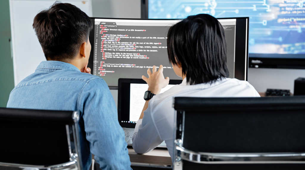 two programmers sit with their backs to the camera as they both examine a coding language on a shared computer screen