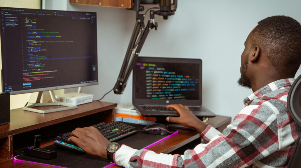 a young man of color sits in front of dual computer screens at a desk, looking at coding language and typing on his keyboard