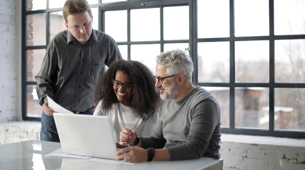 three people of different ages and backgrounds sit together at a conference desk viewing a laptop screen together