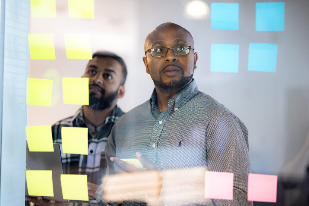 two men stand in front of a glass wall with assorted sticky notes attached, in deep concentration