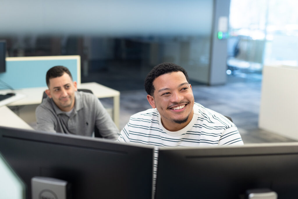 two men sit in front of a computer station looking at a screen together and smiling