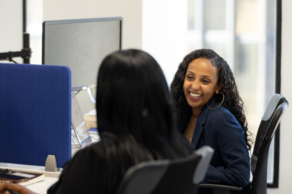 two women sit side by side in front of computer screens, engaged in conversation