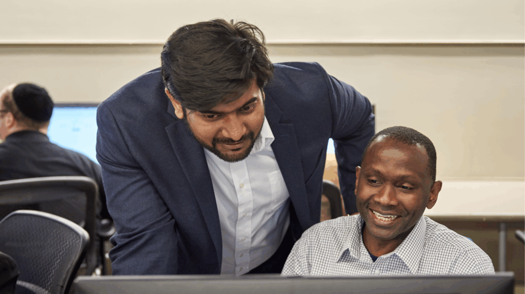 Mentor guiding a technologist over his shoulder, both looking at a computer screen