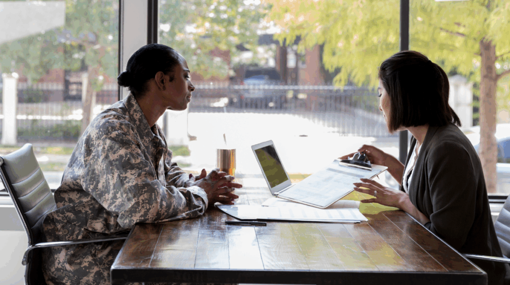 two individuals sit across from one another at a desk, one is dressed in a military uniform