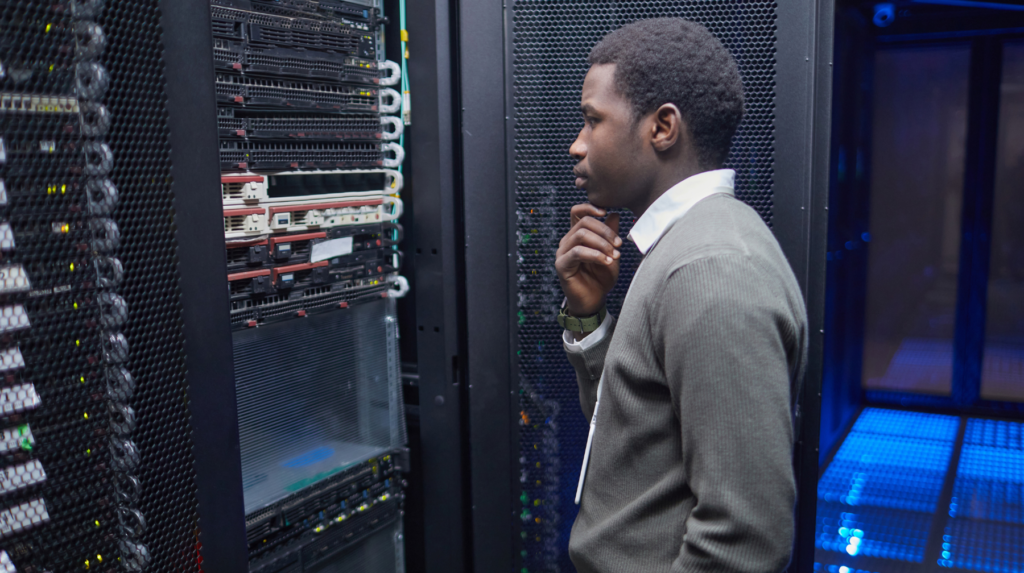 African American technician standing near server racks in a data center, examining equipment and ensuring optimal performance